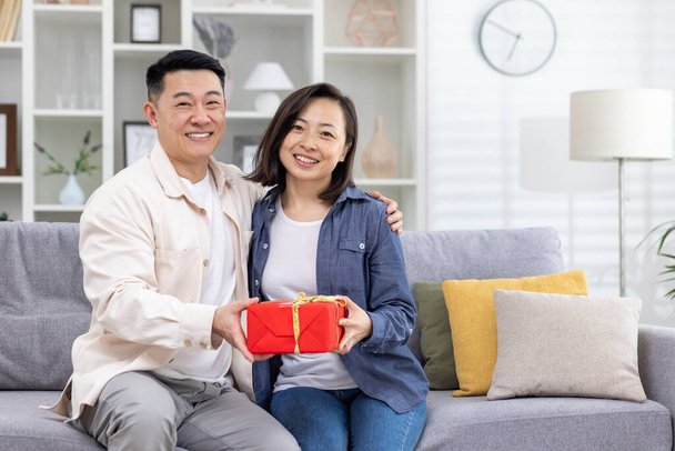 Een jong Aziatisch echtpaar, een man en een vrouw, zitten thuis op de bank en houden een geschenkdoos in hun handen. Lachend en gelukkig kijkend naar de camera. - Foto, afbeelding