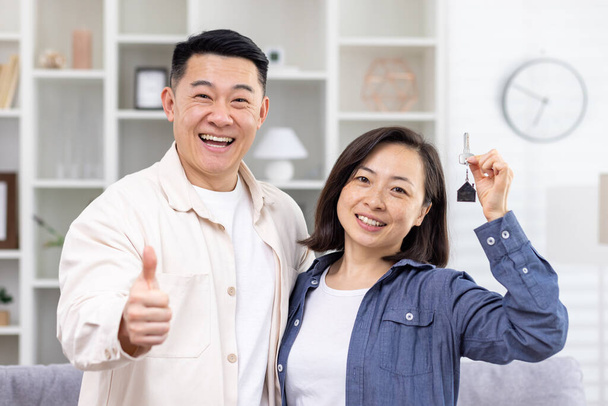 Happy young Asian couple bought an apartment, a house. They stand hugging and smiling at the camera. The man shows super with his finger, the woman holds the keys in her hand. - Foto, afbeelding