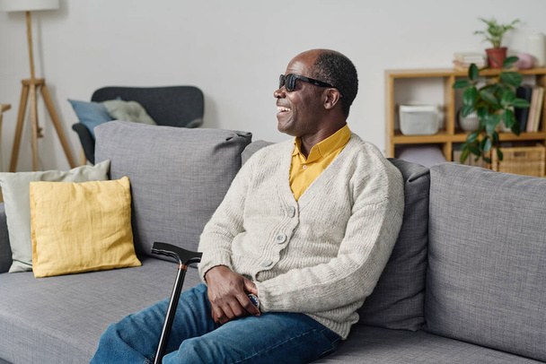 Afričan slepec v tmavých brýlích sedí na pohovce v obývacím pokoji a směje se - Fotografie, Obrázek