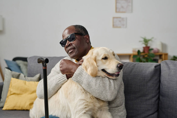 Africano homem sênior em óculos com má visão abraçando seu cão guia individual enquanto sentado no sofá em casa - Foto, Imagem