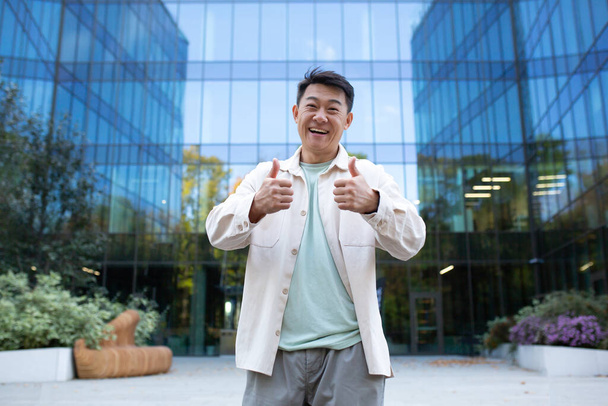 Portrait of young asian male designer, freelancer, businessman standing outside near office center. He stands confidently and looks into the camera with a smile, shows super with his fingers. - 写真・画像