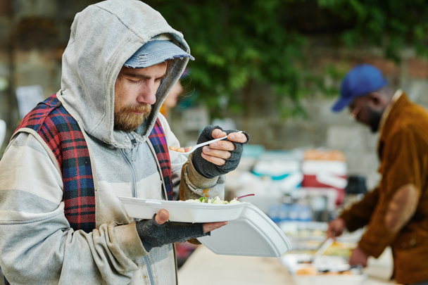 Άστεγος άντρας με ζεστά σχισμένα ρούχα τρώει φαγητό σε εξωτερικούς χώρους δίνοντας από εθελοντές κατά τη διάρκεια φιλανθρωπίας - Φωτογραφία, εικόνα