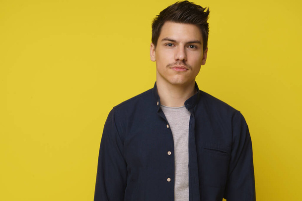 20s years old handsome young man in casual deep blue shirt standing against yellow background - Foto, afbeelding