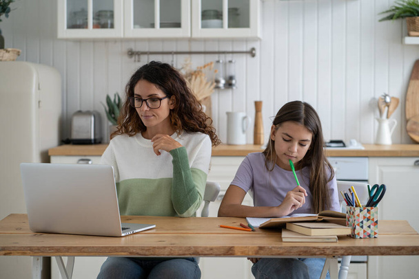 Przemyślana kobieta freelancer patrząc ekran odległej pracy w laptopie siedzi przy stole w kuchni z córką przygotować się do egzaminu. Zamyślona nastolatka z ołówkiem rozmyślać rozwiązanie, czyta książki, co zadanie szkoły - Zdjęcie, obraz