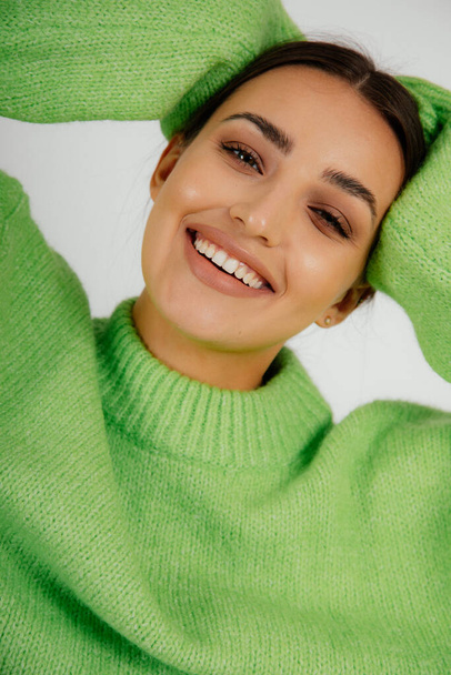Zamknij pionowe zęby uśmiechnięta brunetka z makijażem w zielonym stroju z rękami na głowie pozowanie i patrząc na aparat fotograficzny na białym tle studio. Usługi dentystyczne i zdrowe wybielanie zębów - Zdjęcie, obraz
