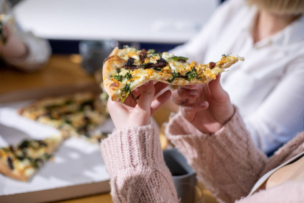 Cropped photo of woman sitting at table among friends relatives , holding piece of vegetable pizza, enjoying food, relaxing at home. Celebration, birthday, fast food, party, togetherness. Soft focus. - Photo, Image