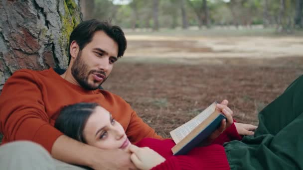 Ihana huoleton pari makaa puistossa kirjan lähikuva. Viehättävä parrakas mies lukee ääneen kauniille naiselle. Rauhallinen nainen rentouttava poikaystävä rinnassa. Onnellinen nuori pari viettää aikaa yhdessä metsässä - Materiaali, video
