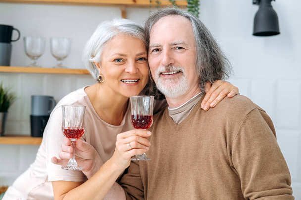 Heureux conjoints caucasiens âgés, passer du temps ensemble dans la cuisine à la maison, tenir des verres de vin dans leurs mains, célébrer une date importante, anniversaire, regarder la caméra, sourire joyeusement - Photo, image