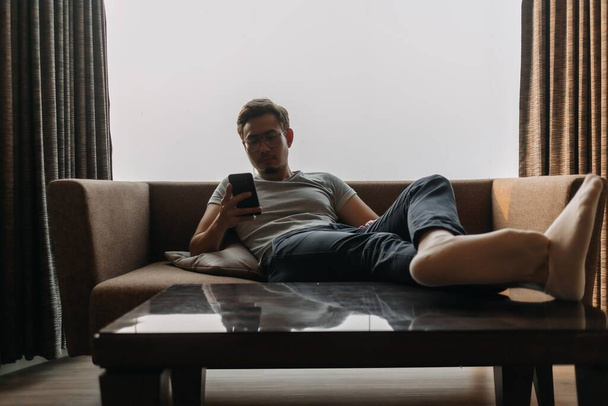 Asiatico uomo si trova sul divano utilizzando il telefono con i piedi sul tavolo. Concetto di modo - Foto, immagini