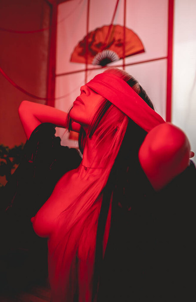 The art of eroticism and bondage: young, slim and sexy woman with only a little black kimono putting on a blindfold, in a beautiful red-lit traditional japanese room - Photo, Image