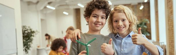 Our invention. Portrait of two happy boys showing thumbs up while holding mechanical toy they assembled on crafting classes. Kids posing for camera at STEM lesson. Education, technology concept - Φωτογραφία, εικόνα