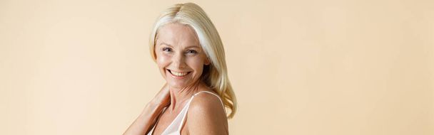 Portrét šťastné dospělé ženy s blond vlasy v bílém spodním prádle se usmívá na kameru, pózuje izolované přes béžové pozadí. Krása, koncepce sebepřijatelnosti - Fotografie, Obrázek