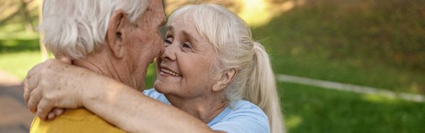Lachende volwassen vrouw met echtgenoot in sportkleding knuffel staan in het groene park op de zomerdag. Actieve levensstijl en gezondheidszorg - Foto, afbeelding