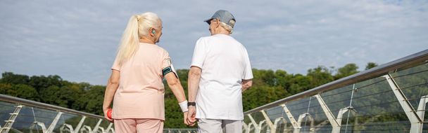 Pozytywny starszy kobieta i mężczyzna chodzić trzymając się za ręce wzdłuż kładki po treningu w letni wieczór widok z tyłu. Aktywny tryb życia i opieka zdrowotna - Zdjęcie, obraz