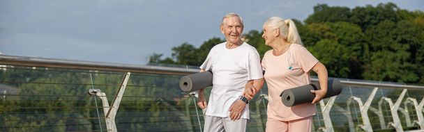 Glückliche Seniorinnen und Senioren mit Rollmatten, die für das Training bereit stehen, spazieren bei Sonnenuntergang über die Fußgängerbrücke. Aktiver Lebensstil und Gesundheitsfürsorge - Foto, Bild
