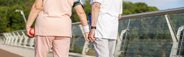 Ηλικιωμένο ζευγάρι σε φόρμες με μπουκάλια βόλτες ενώνει τα χέρια κατά μήκος πεζογέφυρα το καλοκαίρι βράδυ closeup πίσω όψη. Ενεργός τρόπος ζωής και υγειονομική περίθαλψη - Φωτογραφία, εικόνα