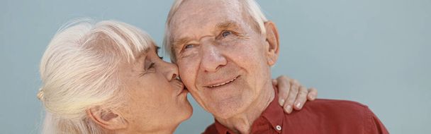 Blonde mature woman kisses loving man partner standing on light grey background in studio. Love and family relationship - Photo, image