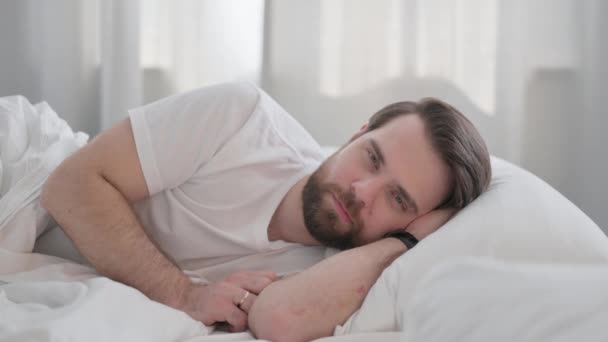 glimlachende jonge volwassen man liggend in bed op kant - Video