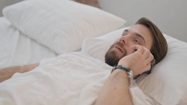 Mladý dospělý muž mluví na telefonu v posteli - Fotografie, Obrázek