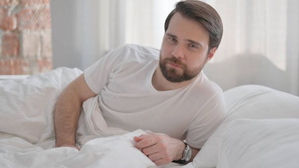 Serious Young Adult Man Lying on Elbow in Bed Looking at Camera - Photo, image