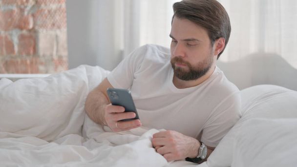 Jonge volwassen man liggend op de elleboog in bed en met behulp van telefoon - Foto, afbeelding