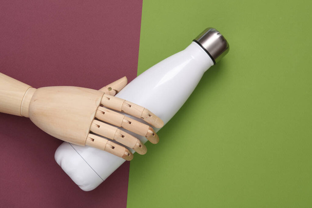 Drewniana dłoń trzymająca białą metalową butelkę termalną do wody na kolorowym tle. Butelka z izolacją próżniową ze stali nierdzewnej - Zdjęcie, obraz