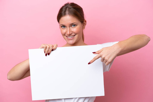 Young caucasian woman isolated on pink background holding an empty placard with happy expression and pointing it - Fotografie, Obrázek