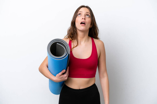 Young sport caucasian woman going to yoga classes while holding a mat isolated on white background looking up and with surprised expression - Photo, image