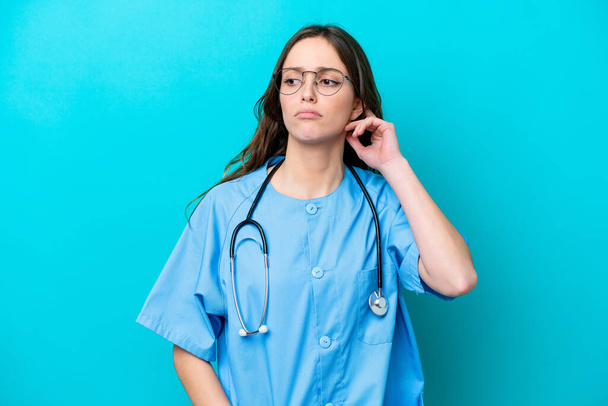 Young surgeon doctor woman isolated on blue background having doubts - 写真・画像