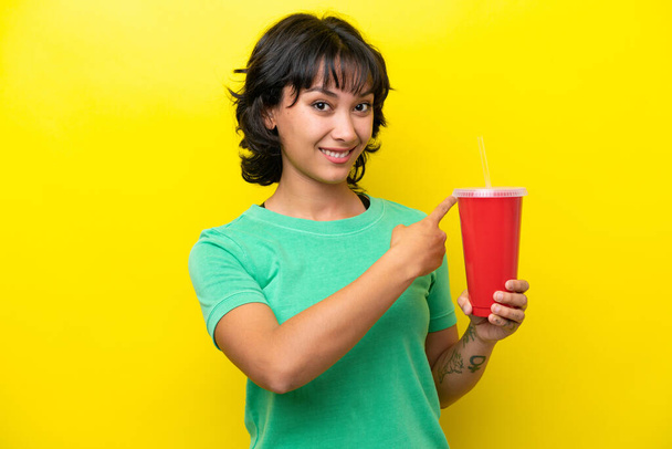 Young Argentinian woman holding a soda isolated on yellow background pointing back - Foto, Bild