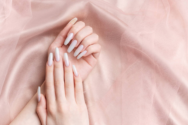 Egy nő manikűrös kezei, a körmök fehér színűre vannak festve. Rózsaszín selyem háttér - Fotó, kép