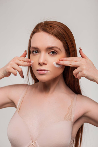 beautiful red-haired girl applies a cream on her face and moisturizes her skin on a white background in the studio - Photo, Image