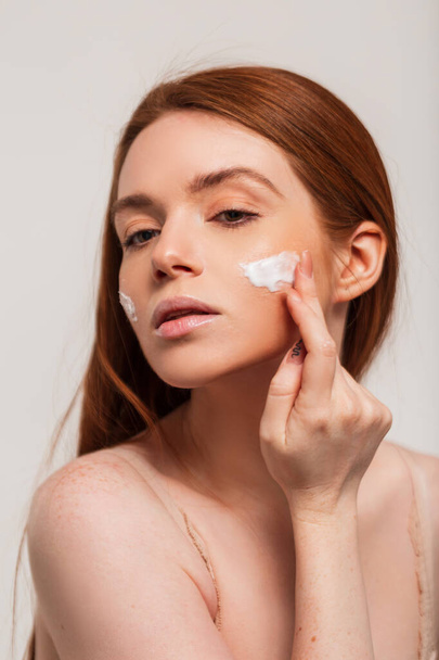 Beautiful fresh young redhead woman with red hair applies a cream on her face and does a skin moisturizing. Beauty, freshness and skin care - Fotó, kép