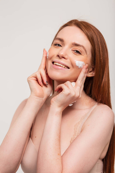 Šťastná krásná mladá žena model s přírodními zrzavými vlasy a roztomilý úsměv aplikuje krém na obličej kůže ve studiu na bílém pozadí. Krása a péče o pleť - Fotografie, Obrázek