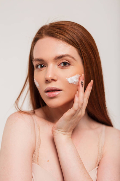 Beautiful young happy redhead fresh lady applying moisturizer cream on face skin in studio. Beauty and skin care - Photo, Image