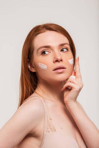 Roodharige mooie frisse jonge vrouw brengt crème aan op de huid van het gezicht in de studio op een witte achtergrond. Schoonheid en huidverzorging - Foto, afbeelding