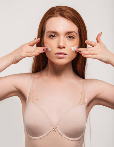 Beautiful fashion model woman with red hair and fresh clean skin in a beige bra applies a cream on her face in the studio. Beauty and skin care - Photo, Image
