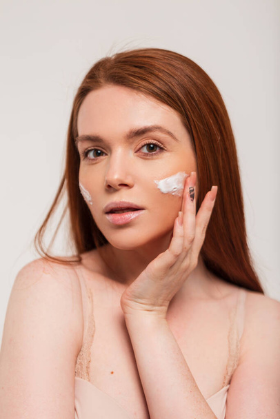 Beautiful young happy redhead fresh lady applying moisturizer cream on face skin in studio. Beauty and skin care - Fotó, kép