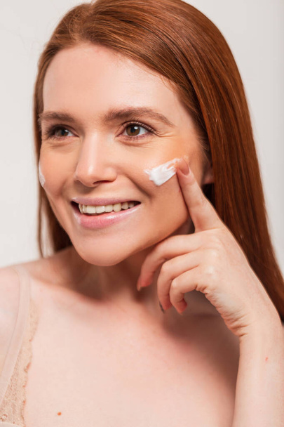 Happy beautiful young redhead woman applying cream on face skin and moisturizing in studio. Skin care and beauty - Photo, Image