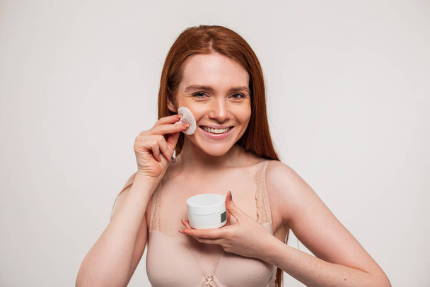 Beautiful happy young fresh redhead with smile girl is holding a cream jar and wiping her face with a cotton pad in the studio. Beauty and skin care - Photo, Image
