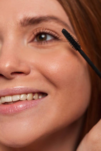 Beautiful happy redheaded woman model with a cute smile paints her lashes with mascara, closeup. Beauty and makeup - Valokuva, kuva