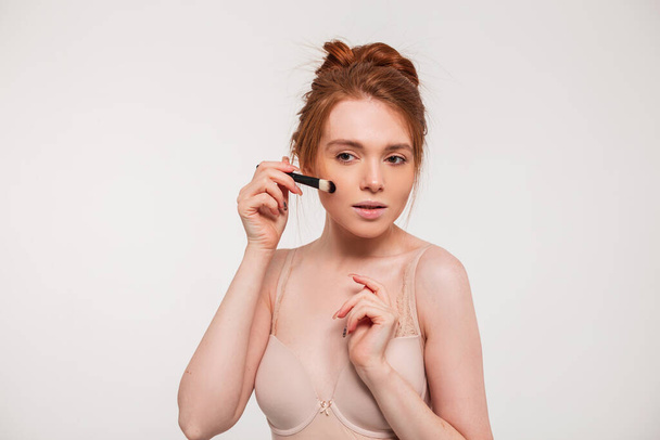 Beautiful cute fresh redhead girl in beige bra with a brush makes up on a white background. Beauty and skin care - Photo, Image