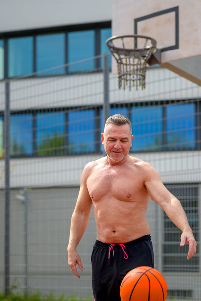 Hombre sin camisa de mediana edad en pantalones cortos deportivos negros gotea y mirando hacia abajo a la pelota de baloncesto en un campo de deportes - Foto, Imagen