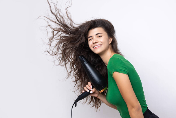 A cute young girl dressed in a green top dries her beautiful long silky hair with a hair dryer against a white background. - Fotoğraf, Görsel