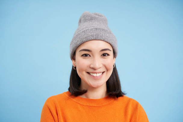Gros plan portrait de souriante femme asiatique au chapeau chaud, regardant heureux et mignon à la caméra, a la peau naturelle claire, se tient sur fond bleu. - Photo, image