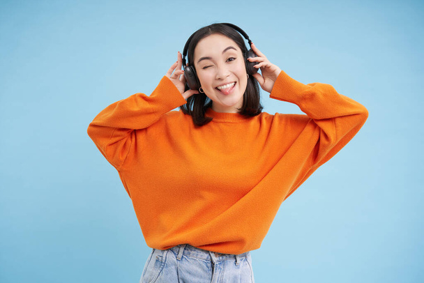 Mulher chinesa feliz em fones de ouvido, ouve música, gosta de música favorita em sua lista de reprodução, fica sobre fundo azul. Espaço de cópia - Foto, Imagem