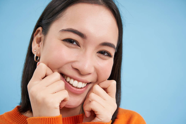 Schönheit und Hautpflege. Nahaufnahme Porträt einer glücklich lächelnden Japanerin, berührt ihre klare, leuchtende Haut, ihr natürliches gesundes Gesicht, steht vor blauem Hintergrund. - Foto, Bild