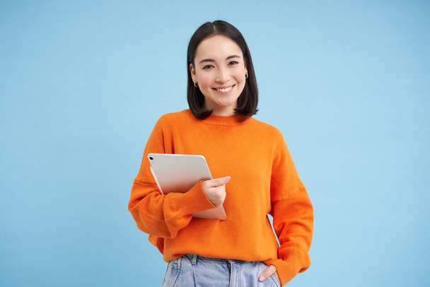 Smiling modern woman with tablet, looking confident, student with her gadget, standing over blue background. - Foto, Imagem