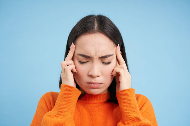 Close up portrait of asian woman with migraine, touches her head and massaging temples, has headache, stands over blue background. - Foto, Imagen