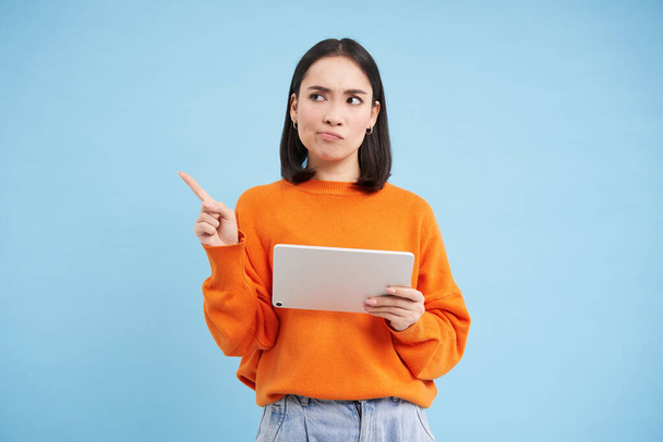 Portrait of korean woman with digital tablet, pointing at upper left corner with confused, frowning face, standing over blue background. - Foto, Bild